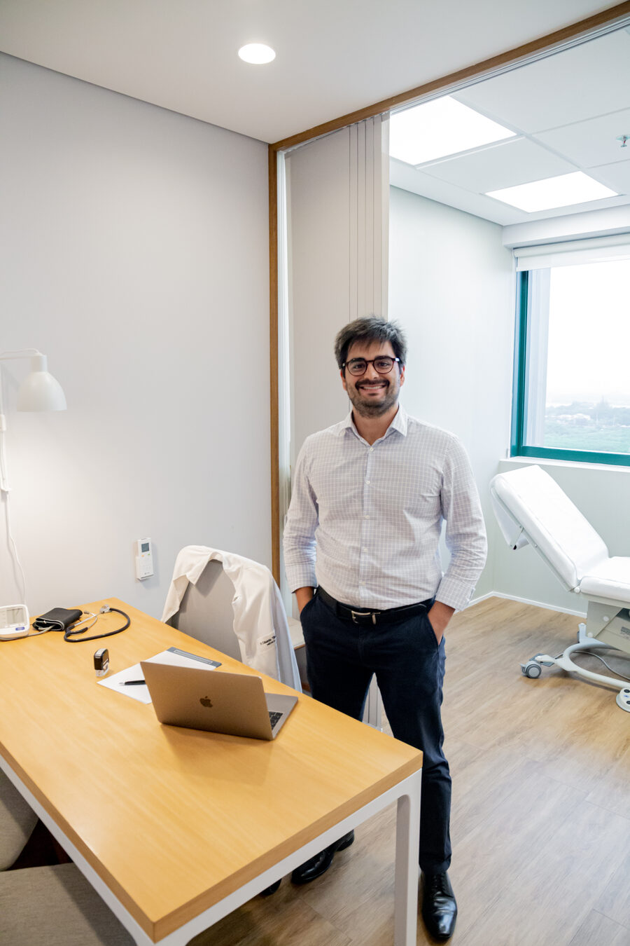 Dr. Eduardo Tiburcio no consultório, ao lado da mesa e da maca de exame